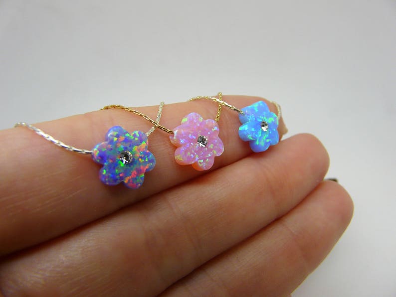 Puede incluir: Tres collares con colgantes de &oacute;palo en forma de flor en azul, rosa y celeste. Cada colgante tiene una peque&ntilde;a piedra central brillante y est&aacute; ensartado en una delicada cadena. Los collares se muestran sobre un fondo claro.