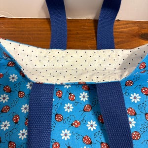 May include: A blue tote bag with navy blue handles. The bag's exterior features a pattern of red ladybugs and white daisies on a light blue background. The interior lining is white with small black polka dots.
