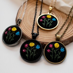 May include: Four necklaces with round pendants featuring embroidered floral designs. The pendants are black with yellow, blue, and pink flowers. The necklaces have silver, black, gold, and bronze chains.