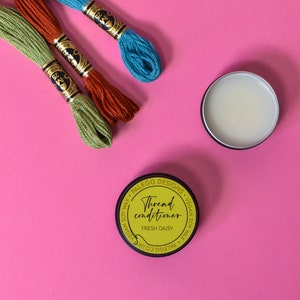 May include: A small yellow jar with a black lid and a label that reads "Thread conditioner Fresh Daisy" on a pink surface. Three spools of embroidery floss in green, orange, and blue are also on the surface.