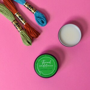May include: A green jar of thread conditioner with the text "Thread conditioner Coconut Lime" on the label. Three spools of embroidery floss in green, orange, and blue are on a pink surface.