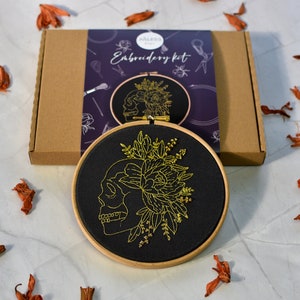 May include: Embroidery kit featuring a black fabric hoop with a gold embroidered skull with floral details. The kit includes all the necessary materials to complete the project. The text "Embroidery Kit" is printed on the box.