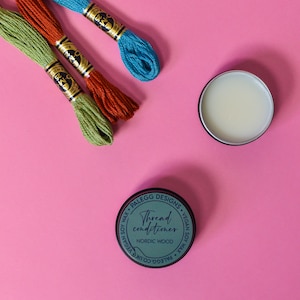 May include: Three spools of embroidery floss in green, orange, and blue, with a small tin of thread conditioner labeled "Thread conditioner, Nordic Wood, Vegan Soy Wax" on a pink background.