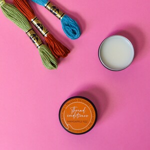May include: Three spools of embroidery floss in green, orange, and blue, with a small jar of thread conditioner labeled "Thread conditioner, Mangapple Fizz" on a pink background.