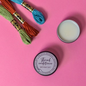 May include: A small tin of thread conditioner with the label "Thread conditioner" and "Birthday Suit" in white text on a purple background. The tin is surrounded by three spools of embroidery floss in green, orange, and blue.
