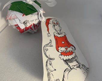 May include: A Christmas ornament featuring a Dr. Seuss character illustration on a white tag. The ornament is paired with a paper ball with red and white accents and a green top, tied with a white ribbon.