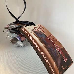 May include: A decorative ornament shaped like a pinecone, made from layered paper. A black ribbon is attached for hanging. A rectangular tag with the text "New Miserable Experience" and a photo is attached.