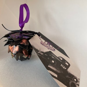 May include: A unique, handmade ornament with a purple ribbon for hanging. The ornament is made of folded black and purple paper with images of a person. A tag with the text "Aerosmith" and a black and white image of a car is attached.