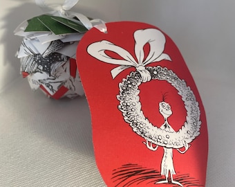 May include: A Christmas ornament featuring a red tag with a black and white illustration of a character holding a wreath. The ornament is attached to a paper ball with a white ribbon. The paper ball is made of pages with black and white illustrations and green paper.