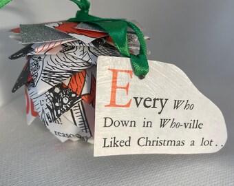 May include: A handmade Christmas ornament shaped like a pinecone, crafted from book pages with red and silver accents. A white tag with the text "Every Who Down in Whoville Liked Christmas a lot.." is attached with a green ribbon.