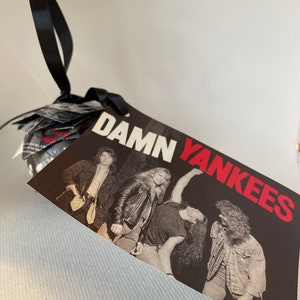 May include: A black tag with the words "DAMN YANKEES" in red and white, featuring a black and white photo of a band. The tag is attached to a bundle with a black ribbon.