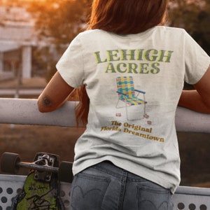 Puede incluir: Una camiseta blanca con el texto "LEHIGH ACRES" y un gráfico de una silla plegable. El texto "The Original Florida Dreamtown" está debajo del gráfico de la silla.