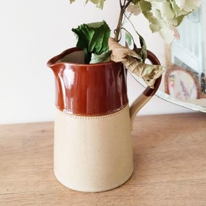 May include: A two-toned ceramic pitcher with a brown top and a cream-colored base. The pitcher is filled with dried hydrangeas and greenery. The pitcher has a handle and a spout. The pitcher is sitting on a wooden surface.
