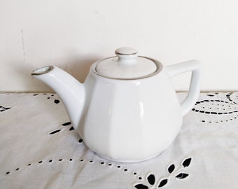 May include: A white ceramic teapot with a lid and handle, set on a white lace tablecloth. The teapot has a geometric design and a silver rim. Two gold-coloured bird decorations are mounted on the wall in the background.