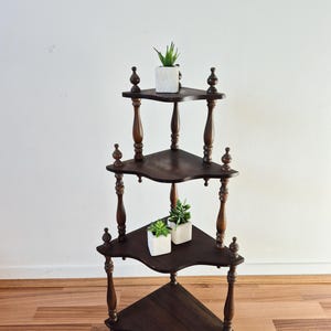 May include: A dark brown wooden corner shelf unit with four tiers, featuring turned spindle supports and decorative finials. The shelves have curved edges. Three small white square pots containing green artificial succulents are placed on the shelves.
