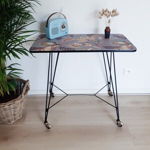 Peut inclure: Une petite table d'appoint en métal noir avec un plateau à motif floral. La table a quatre pieds et des roulettes. Une radio bleue et un vase avec des fleurs séchées sont sur la table.