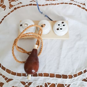 Peut inclure: Une prise électrique vintage en porcelaine blanche avec trois interrupteurs ronds et une chaînette de tirage brune avec une ampoule en bois. La prise est sur une nappe en dentelle blanche.