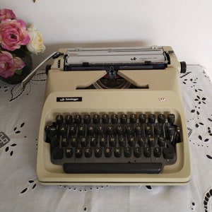 May include: Cream-colored Scheidegger International 2000 typewriter with black keys. The keyboard layout is AZERTY. The typewriter sits on a white lace tablecloth. A gold-colored bird decoration and a bouquet of pink and white flowers are in the background.