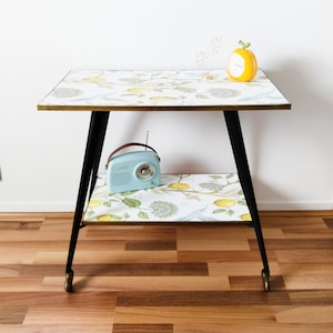 Può includere: Un carrello da servizio vintage a due livelli con gambe nere e dettagli dorati. Il carrello ha un piano superiore bianco con un motivo floreale e una radio blu chiaro sullo scaffale inferiore.