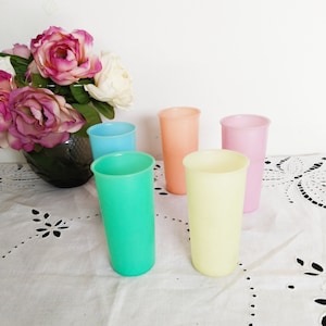 May include: Five pastel colored drinking glasses. The glasses are light blue, green, yellow, orange, and pink. The glasses are arranged in a row on a white tablecloth.