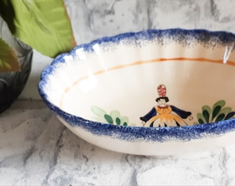 May include: A white oval ceramic bowl with a blue and orange rim and a hand-painted design of a woman in a yellow dress and blue hat. The bowl is decorated with green leaves and flowers.