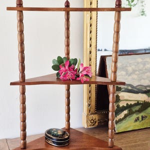 May include: A three-tiered wooden corner shelf featuring turned spindle supports and triangular shelves. The unit has a medium brown finish. A small black oval box with a floral design sits on the bottom shelf, and a sprig of pink flowers rests on the middle shelf.