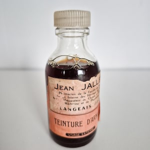 May include: A small, clear glass bottle with a light brown liquid inside. The bottle has a beige cap and a label with French text, including "TEINTURE D'ARN" and "USAGE EXTERNE".