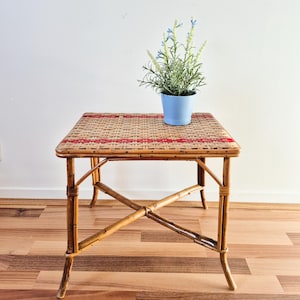 May include: A square side table constructed from natural brown rattan with a woven top featuring horizontal red accent stripes. The table has four legs connected by a central X-shaped support brace. A light blue pot with green foliage sits on top.