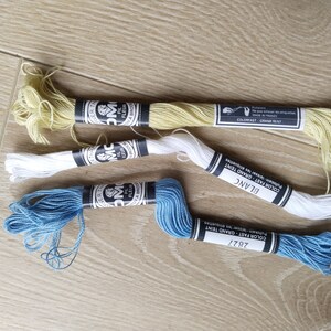 May include: Three spools of embroidery floss in different colors: yellow, white, and blue. The spools are labeled with the brand name "DMC" and the color names "Grand Teint", "Blanc", and "2827".
