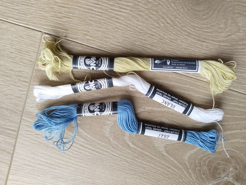 May include: Three spools of embroidery floss in different colors: yellow, white, and blue. The spools are labeled with the brand name "DMC" and the color names "Grand Teint", "Blanc", and "2827".