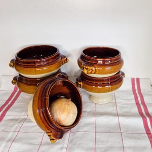 Peut inclure: Quatre bols à soupe vintage en céramique marron et beige avec des poignées sont empilés par paires. Un bol est ouvert, révélant un gros oignon. Les bols ont un design classique et rustique, parfait pour servir de la soupe ou du ragoût.