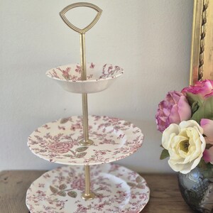 Peut inclure: Présentoir à trois niveaux avec une poignée et un support dorés. Les assiettes et le bol présentent un motif floral dans les tons de rose et de vert sur fond blanc. Le présentoir est posé sur une surface en bois.