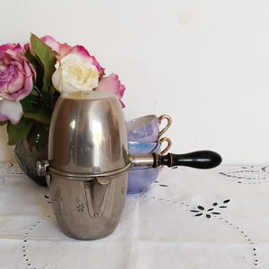 Può includere: Una teiera d'argento con un manico nero e un design decorativo. La teiera è appoggiata su una tovaglia bianca con un motivo floreale.