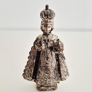 May include: A small, silver-coloured statue of a crowned figure, possibly religious. The figure wears a detailed robe and holds objects in each hand. The statue has intricate detailing and stands on a small base. The background is plain white.