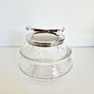 Peut inclure: Un pot en verre transparent craquelé, surmonté d'un couvercle et d'une poignée en métal argenté. Le pot a une forme arrondie avec un design à deux niveaux. Le couvercle comporte une poignée incurvée avec des extrémités décoratives. Pour le stockage ou l'affichage.