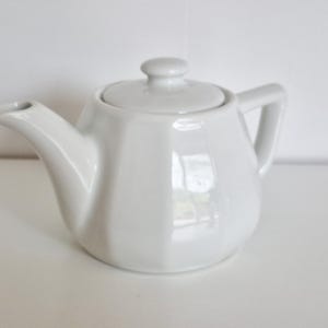 May include: A white ceramic mug with a black handle and the text "But First, Coffee" in a bold, black font. The mug is set against a blurred background, highlighting its clean design.