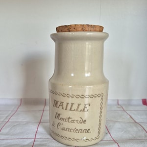 May include: A vintage off-white ceramic jar with a cork stopper. The jar features the text "MAILLE Moutarde à l'ancienne" in brown lettering within a decorative border. The jar is sitting on a white and red checkered surface.
