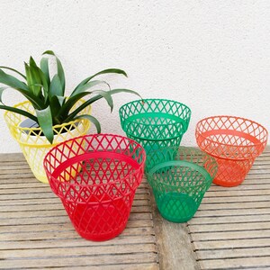 Peut inclure: Quatre pots de fleurs en plastique de différentes couleurs : jaune, vert, rouge et orange. Les pots sont en plastique tressé et présentent un motif en losange. Les pots sont disposés sur une surface en bois.