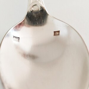 May include: Close-up of a silver spoon with a rounded bowl and a decorative handle. The spoon has a shiny, reflective surface. The bowl of the spoon has two small, square markings with the number "18" visible on one of them.
