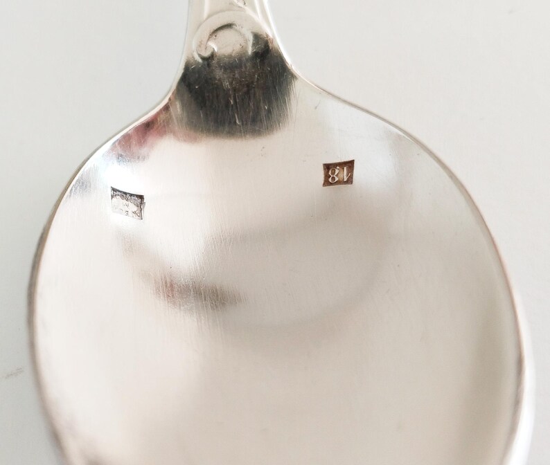 May include: Close-up of a silver spoon with a rounded bowl and a decorative handle. The spoon has a shiny, reflective surface. The bowl of the spoon has two small, square markings with the number "18" visible on one of them.