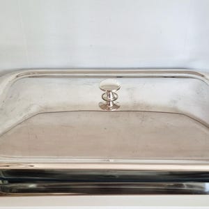 Könnte beinhalten: Eine rechteckige Servierschale in Silberoptik mit passendem Deckel und zwei geschwungenen Seitengriffen. Der Deckel verfügt über einen mittigen Knauf zum Anheben. Die Schale hat eine polierte Metalloberfläche und steht auf einem flachen Tablett.