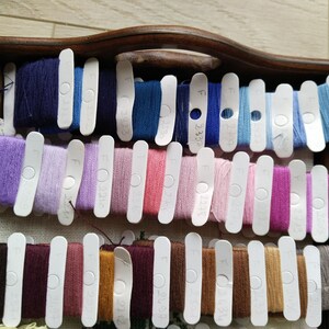 May include: A wooden tray with rows of colorful embroidery floss wound on white plastic bobbins. Each bobbin has a number printed on it, such as "F 00315".