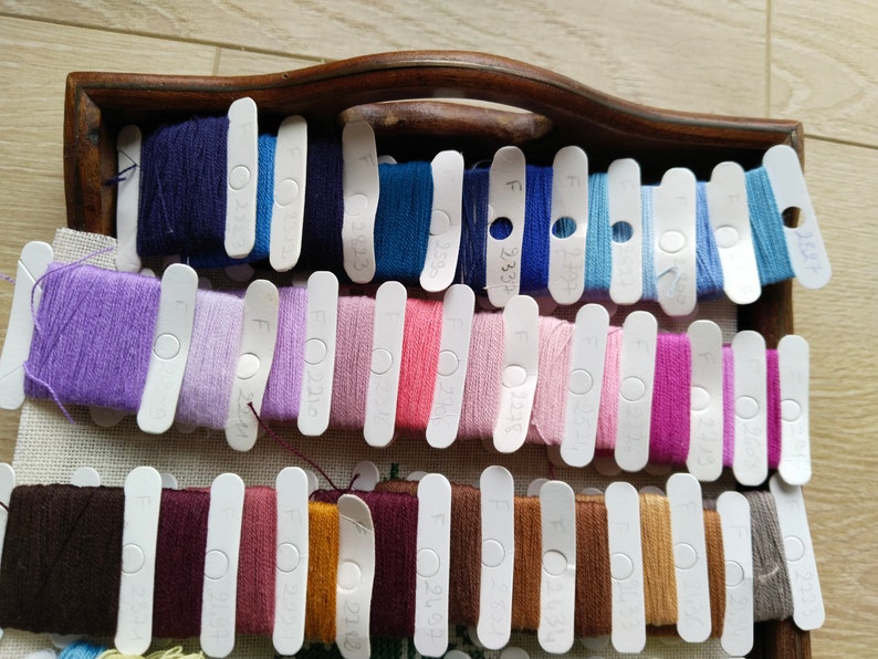 May include: A wooden tray with rows of colorful embroidery floss wound on white plastic bobbins. Each bobbin has a number printed on it, such as "F 00315".