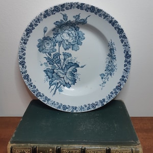May include: A white ceramic plate with a blue floral design. The plate has a scalloped edge and a blue floral pattern around the rim. The centre of the plate features a larger floral design with blue leaves and flowers.