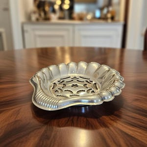 Può includere: Un piatto argentato smerlato con un motivo traforato decorativo al centro. Il piatto ha una forma a conchiglia con un bordo a coste su un lato. Il piatto è probabilmente in metallo e si trova su un tavolo di legno scuro.