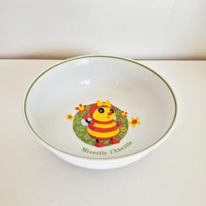 Peut inclure: Un bol en céramique blanche avec un bord vert et une illustration centrale d'une abeille rayée jaune et rouge portant un nœud jaune. Le personnage est entouré de fleurs orange sur un fond circulaire vert avec le texte Mireille l'Abeille.