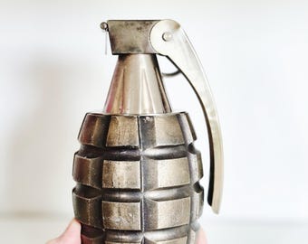 May include: A metallic, bronze-colored grenade-shaped object with a lever and pin. The body has a textured, segmented design. The object is held in a person's hand against a white background. The grenade is a decorative item.