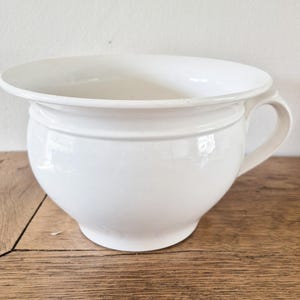 May include: A white ceramic chamber pot with a rounded body, a wide rim, and a curved handle. The pot has a glossy finish and sits on a wooden surface. The design is simple and classic, suitable for vintage or farmhouse decor.