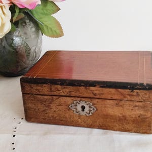 Pode incluir: Uma caixa de madeira retangular com fechadura e buraco da fechadura. A caixa tem um acabamento castanho escuro com um topo castanho mais claro. Um arranjo floral decorativo e livros estão no fundo.