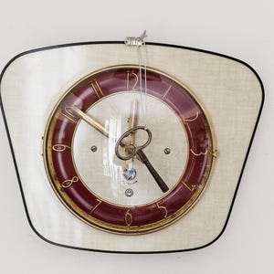 May include: Vintage wall clock with a cream face and a burgundy clock face. It features gold hands and numerals. The clock is encased in a black-edged, uniquely shaped frame. A classic timepiece.
